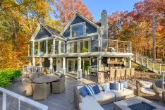 Alt text: "A luxurious two-story house with a large outdoor decking area furnished with sofas and a dining set, surrounded by autumn-colored trees under a clear blue sky."