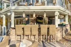 Elegant outdoor patio area with wicker bar chairs, a stone fireplace, and a built-in barbecue grill under a covered deck.