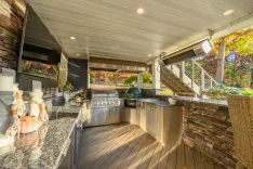 An outdoor kitchen on a deck with stainless steel appliances, a ceramic smoker, stone finishes, and a view of autumn trees.