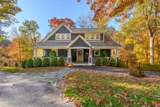 A charming two-story house with a gray exterior and white trim, featuring a covered front porch, autumn foliage, and seasonal decorations.