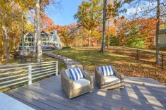 Two wicker chairs with striped pillows on a deck overlooking a scenic backyard with autumn foliage and a large house with a screened porch.