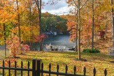 Autumn landscape featuring a lakeside view with colorful trees in red, orange, and yellow hues, a small dock with chairs, and a wrought iron fence in the foreground.