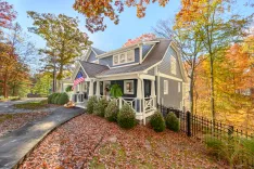 Alt: A charming two-story house with gray siding and white trim surrounded by autumn foliage, featuring a front porch with an American flag and pumpkins by the pathway.