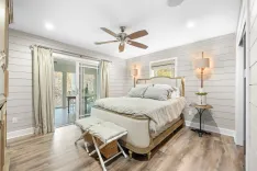 Bright and modern bedroom with a queen-sized bed, two bedside lamps, a ceiling fan, a folding chair with a cushion, sliding glass doors leading to an outdoor area, and shiplap walls.