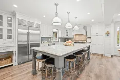 Bright contemporary kitchen interior with white cabinets, marble countertop island, bar stools, stainless steel appliances, and pendant lighting.