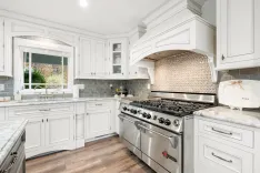 Modern kitchen interior with white cabinetry, stainless steel gas stove and oven, herringbone backsplash, and marble countertops.