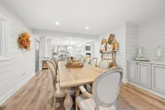 Spacious modern kitchen and dining area with white shiplap walls, wooden dining table, elegant upholstered chairs, and decorative accessories.