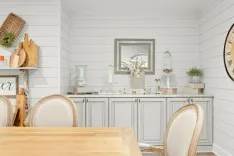 A cozy kitchen interior with white shiplap walls, marble countertops, and farmhouse decor including a wooden table, upholstered chairs, decorative glass jars, and a wall clock.