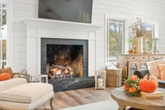 Cozy living room with a lit fireplace, a beige sofa with orange pillows, wooden furniture, autumn decorations, and a view of trees through a window.