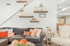 A cozy living room with a brown L-shaped couch adorned with orange and neutral tone pillows, a round wooden coffee table, decorative floating shelves on a white shiplap wall, and a staircase leading to the upper level.