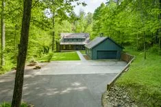 A serene house with a blue garage surrounded by lush greenery in a forest setting with a clear driveway and landscaped lawn.