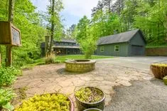 Sunny view of a residential country house with a green exterior, stone courtyard with a fire pit, surrounded by lush trees.