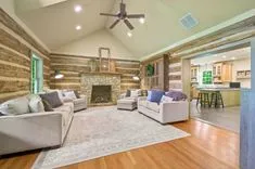 Spacious living room with rustic wood walls and stone fireplace, vaulted ceiling with fan, large sofas, area rug, and adjacent open concept kitchen.