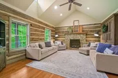 Spacious living room with rustic log walls, a large stone fireplace, vaulted ceiling with fan, hardwood floors, and neutral-colored furniture.