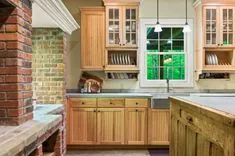 A cozy kitchen interior with wooden cabinetry, countertop, exposed brick column, and a window overlooking green trees.
