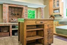 A rustic kitchen interior featuring a brick fireplace, a vintage wooden island with drawers, and wooden cabinetry with a window overlooking greenery.