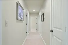 Brightly lit hallway in a home with beige carpet, white walls decorated with framed artwork, multiple closed white doors, and recessed ceiling lighting.