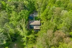 Aerial view of a two-story house with green roofing surrounded by dense trees in a lush green forest clearing.