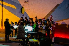 A group of people, including children and adults, wearing festive attire and holding sheet music, singing in a choir beside a lit-up Christmas tree and a piano, against a white brick wall with a painted geometric design.