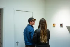 Two people standing in a gallery looking at small artworks on a white wall.