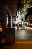 Sidewalk at nighttime with people walking past trees adorned with festive lights, a signboard in the foreground, and streaks of light from a passing vehicle.