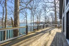 Sunny lakeside wooden deck with railing next to a house, surrounded by bare trees with a clear blue sky.
