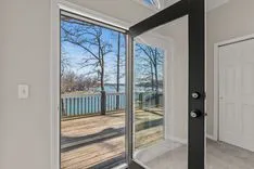 View from a bright room through an open sliding glass door onto a balcony with a view of bare trees and a lake.
