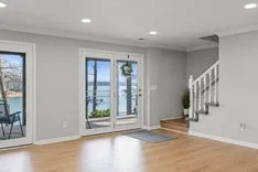 Empty room with hardwood floors, white walls, and glass doors leading to a balcony overlooking a body of water.