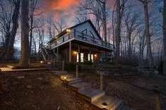 Wooden cabin with lit interior windows at dusk surrounded by bare trees.
