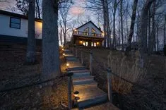 Twilight view of a cozy house with lit windows nestled among bare trees with a lighted stairway leading towards it.
