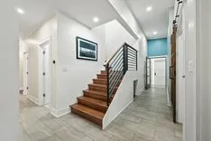Bright modern home interior with a staircase featuring wooden steps and black metal railings, framed artwork on the wall, and tiled flooring.