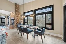 A modern dining room with a wooden table, blue upholstered chairs, large windows, pendant lights, and a stone accent wall.