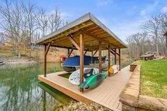 Wooden boat dock with a roof sheltering a speedboat and kayaks on a calm river with trees along the bank.