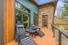 Cozy patio area with two Adirondack chairs and a small table on a wooden deck, adjacent to a house with blue siding and stone accents.