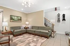 Spacious living room with beige walls, patterned rug, sectional sofa, wooden end tables, lamps, and decorative wall sconces near staircase.
