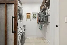 Interior of a modern laundry room with stacked washer and dryer, cabinets, and clothes hanging on the right.