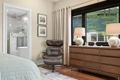 Elegant bedroom interior with a modern leather chair, wooden dresser, table lamps, and framed artwork, with a view of a lush green outdoors through the window.