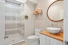 Modern bathroom interior with glass shower stall, white toilet, round mirror, and wooden vanity with vessel sink.