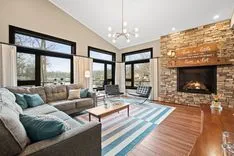 Contemporary living room with large windows, a stone fireplace, hardwood floors, and a blue striped rug.