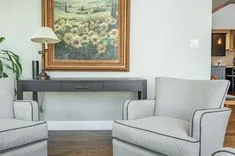 Modern living room with two gray armchairs, a wooden console table, a lamp, and a framed painting of flowers above the table.