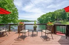 Lakeside deck with patio furniture and red umbrellas overlooking calm water surrounded by green foliage.