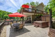 Backyard patio with dining set and red umbrella, adjacent to a hot tub and two-story house with a deck, surrounded by trees on a sunny day.