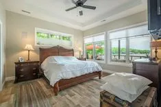 Bright and airy bedroom with a queen-sized bed, wooden furniture, ceiling fan, and large windows showing greenery outside.