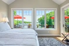 Bright bedroom with a large bed, area rug, and floor-to-ceiling windows overlooking a deck with red umbrellas and a lake view.
