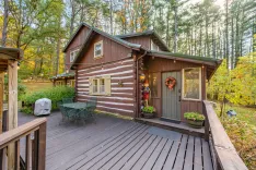 A cozy wooden cabin with a large deck surrounded by lush forest in daylight.