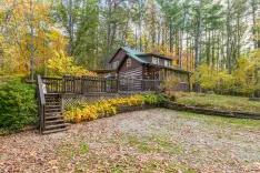 Quaint log cabin surrounded by autumn foliage with a front deck and stone driveway.