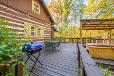 Wooden deck of a house with outdoor furniture, a blue barbecue grill, and a hot tub surrounded by trees in autumn.