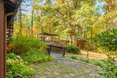 Stone pathway leading to a wooden bridge in a serene autumn forest setting with a cabin in the background and colorful foliage around.