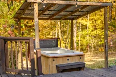 Outdoor wooden hot tub on a deck with a pergola overhead, surrounded by autumn foliage.