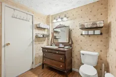 A vintage styled bathroom with patterned wallpaper, wood flooring, a white toilet, vanity cabinet with a mirror and lights overhead, and wall-mounted shelves with towels.